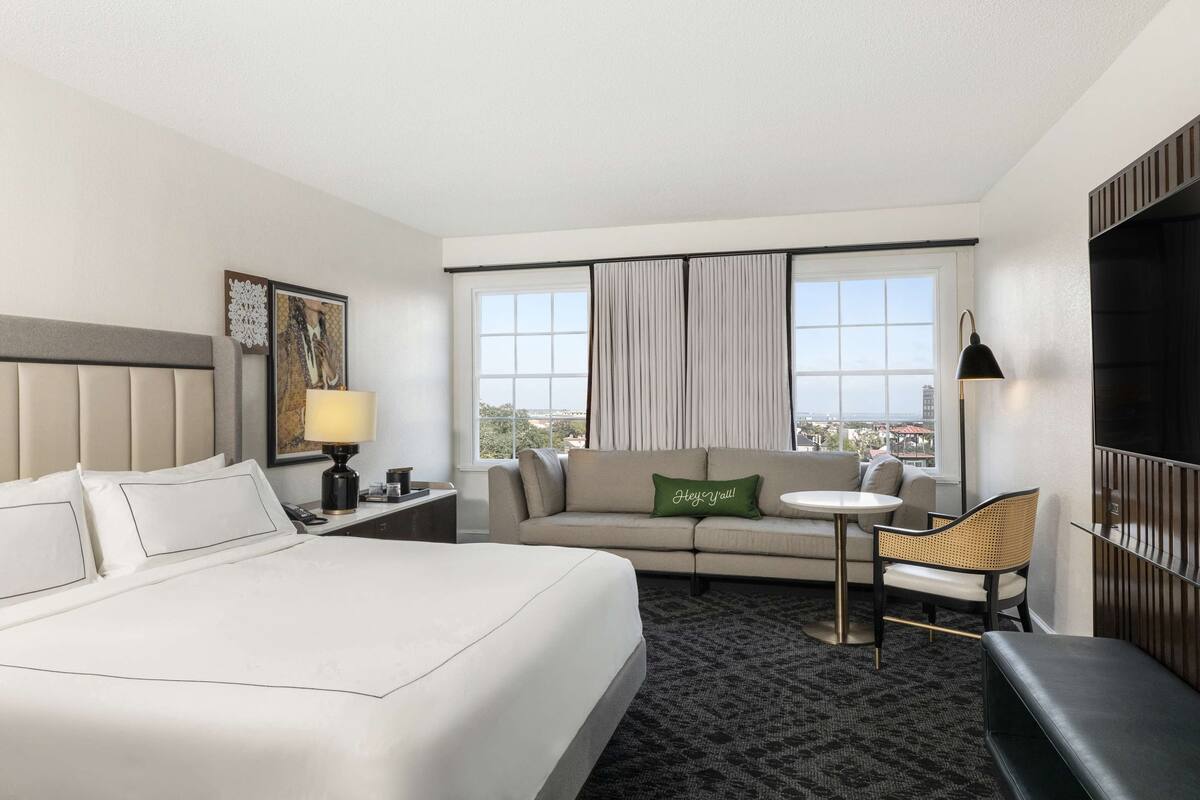 A hotel room with a large bed, a sofa, a small table, and a chair. There is a window with curtains, a lamp, and a painting on the wall.