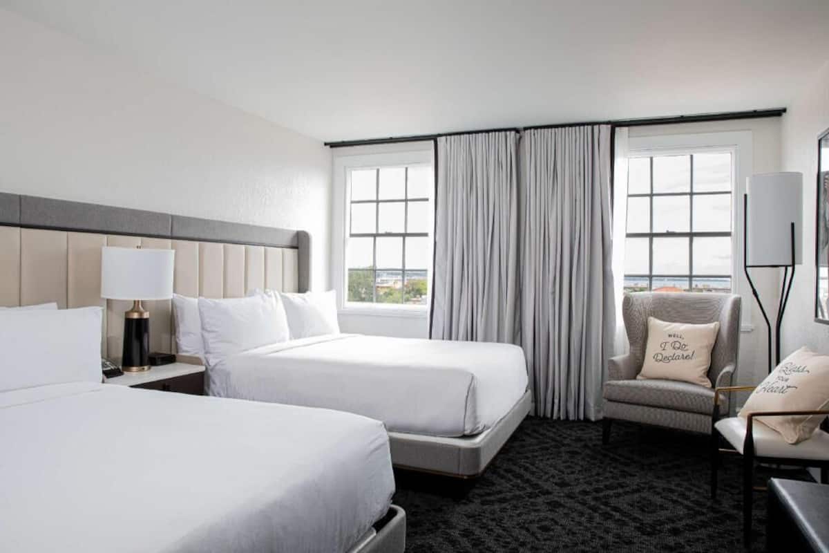 Habitación de hotel con dos camas, una silla, una lámpara y vistas a la ciudad.