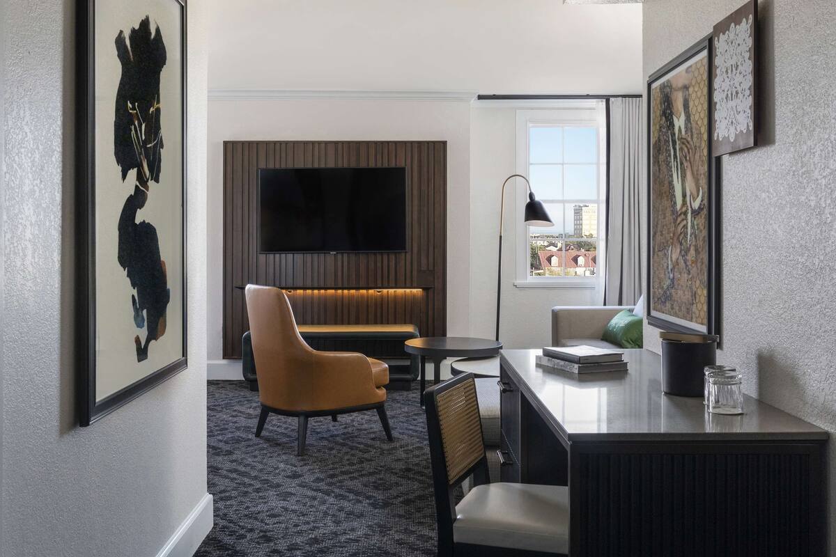 A modern hotel room with a flat-screen TV mounted on a wooden panel wall, a comfortable sofa, a dining table with chairs, and a large window offering a city view.