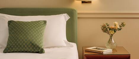 A neatly made bed with a green and white patterned pillow, a bedside table with a vase of flowers, and a lamp.