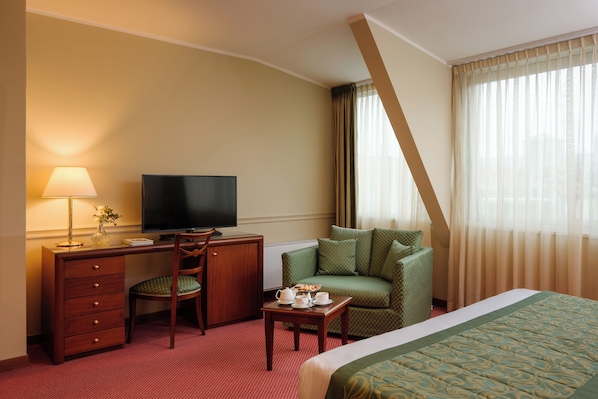 A hotel room with a bed, a desk with a television, a chair, a small table with a cup and saucer, and a sofa.