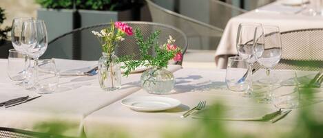 Un área de comedor al aire libre con mesas dispuestas para dos, rodeada de follaje y un edificio al fondo.