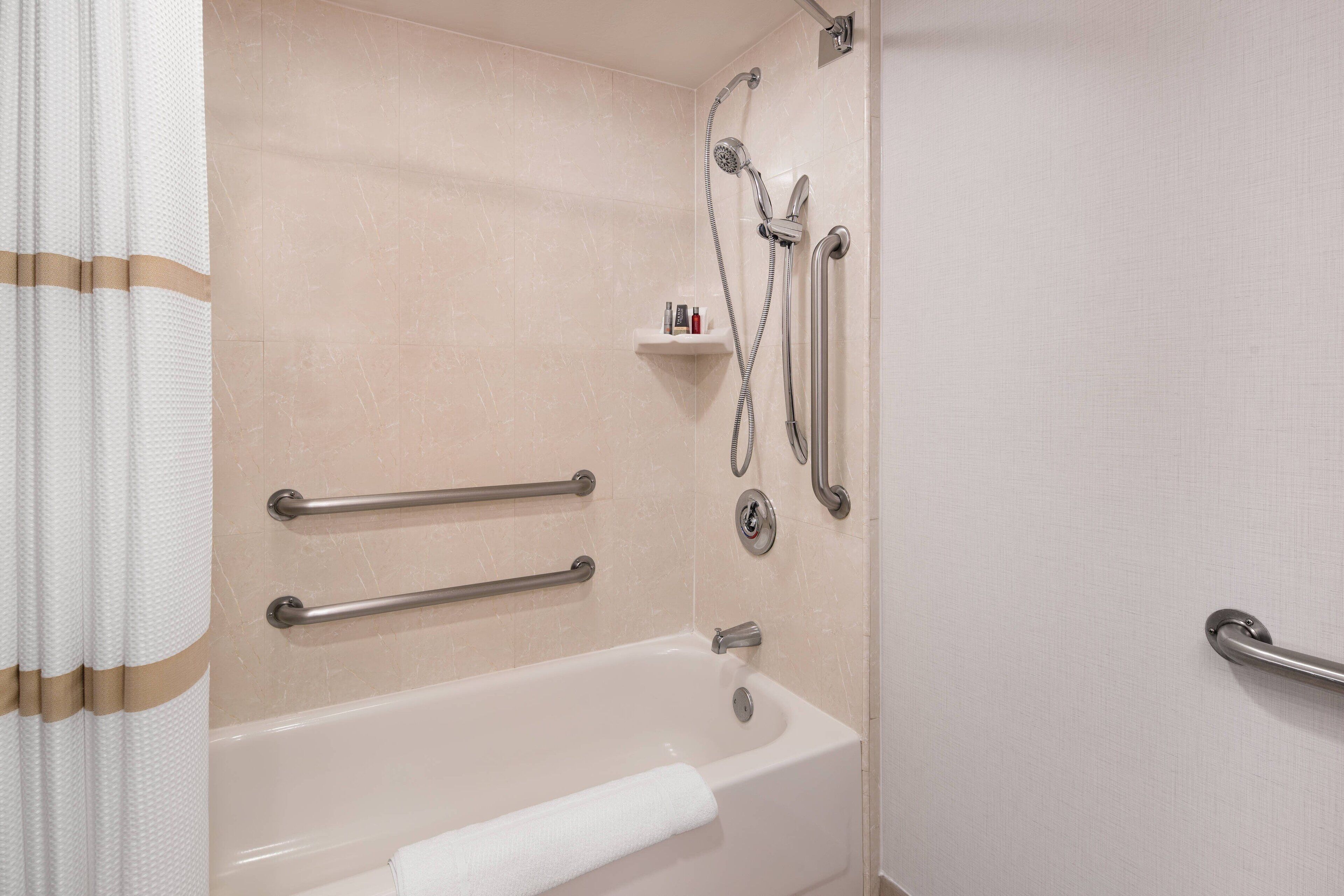 A bathroom with a bathtub, grab bars, a shower with a handheld showerhead, and a shelf with toiletries.