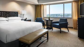 A hotel room with a large bed, a bench, two armchairs, a small table with a plant, a TV, and a window with a city view.