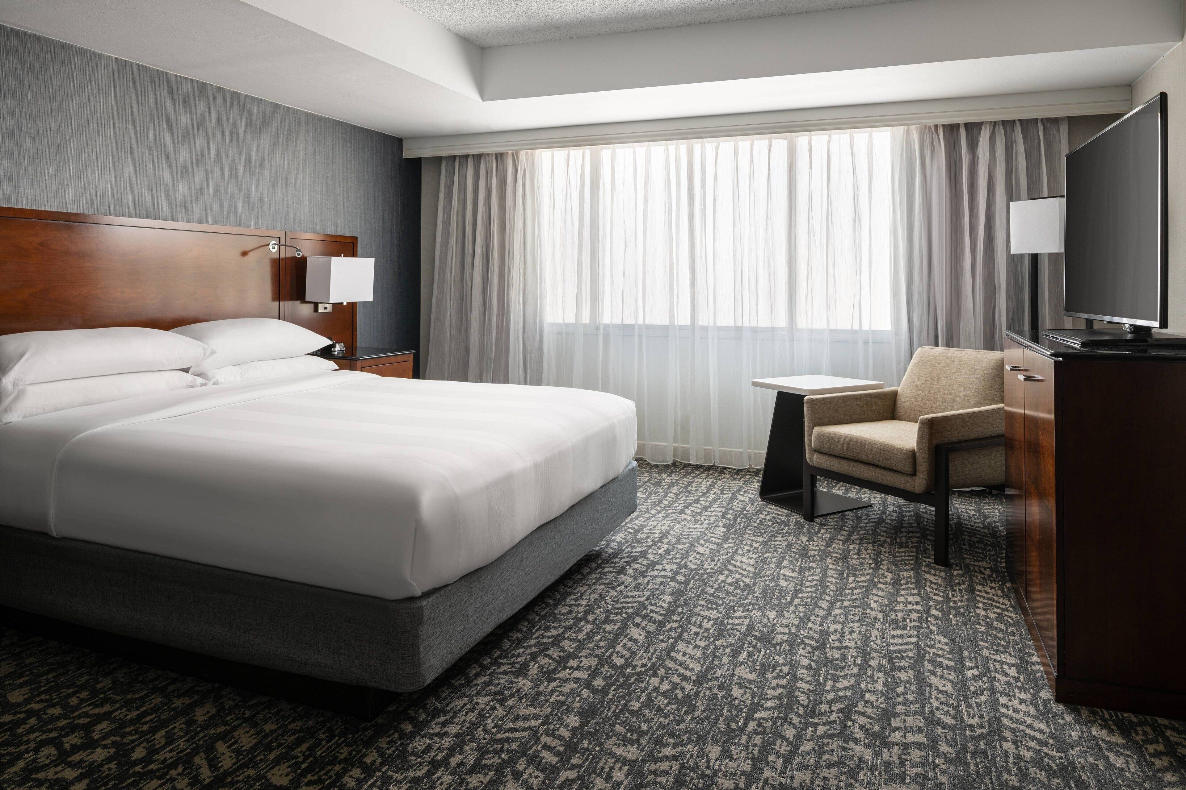 A hotel room with a large bed, a desk, a chair, a TV, and a window with curtains.