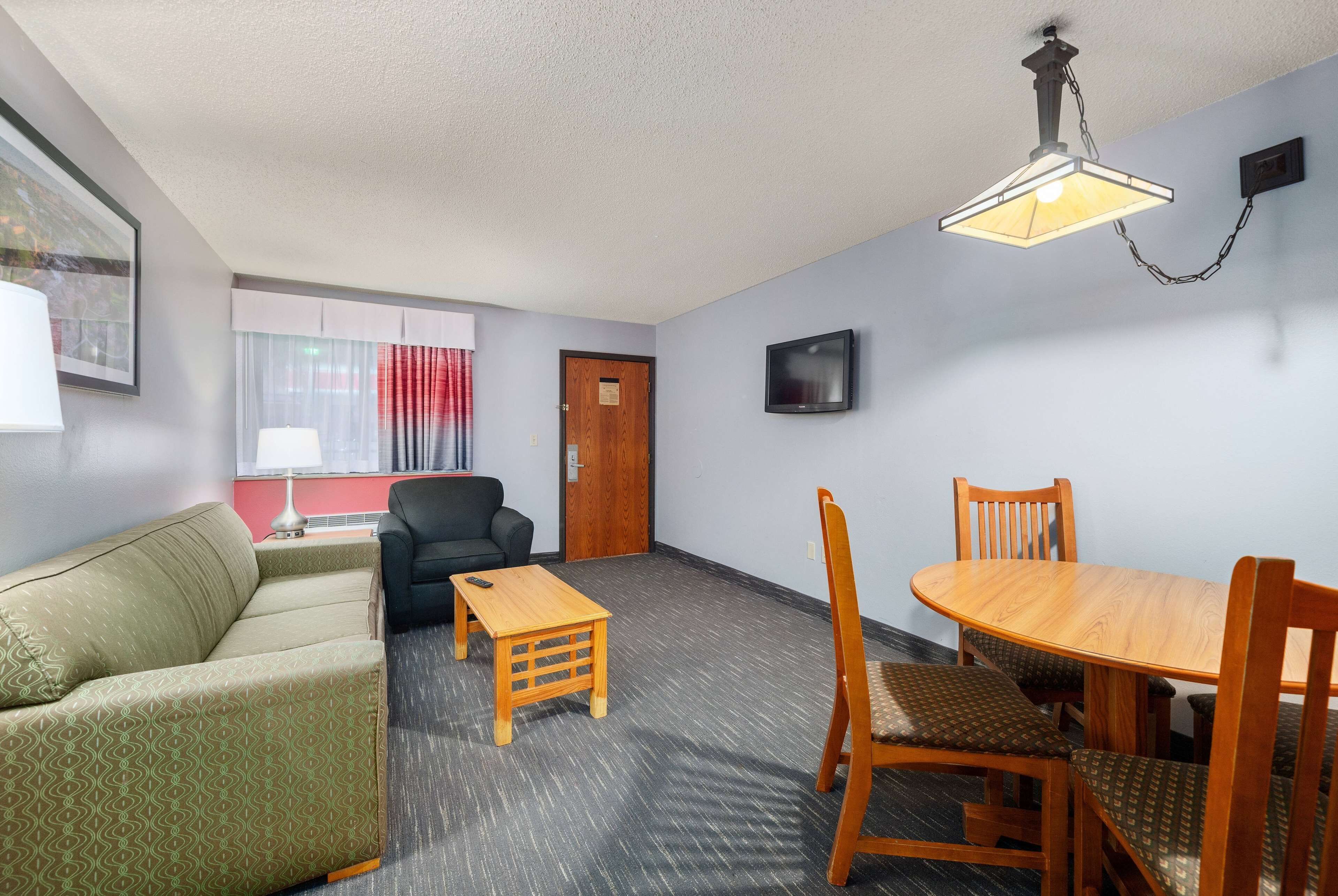 A room with a sofa, a small table, a dining table with chairs, a TV mounted on the wall, and a door.