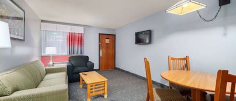 A room with a sofa, a small table, a dining table with chairs, a TV mounted on the wall, and a door.