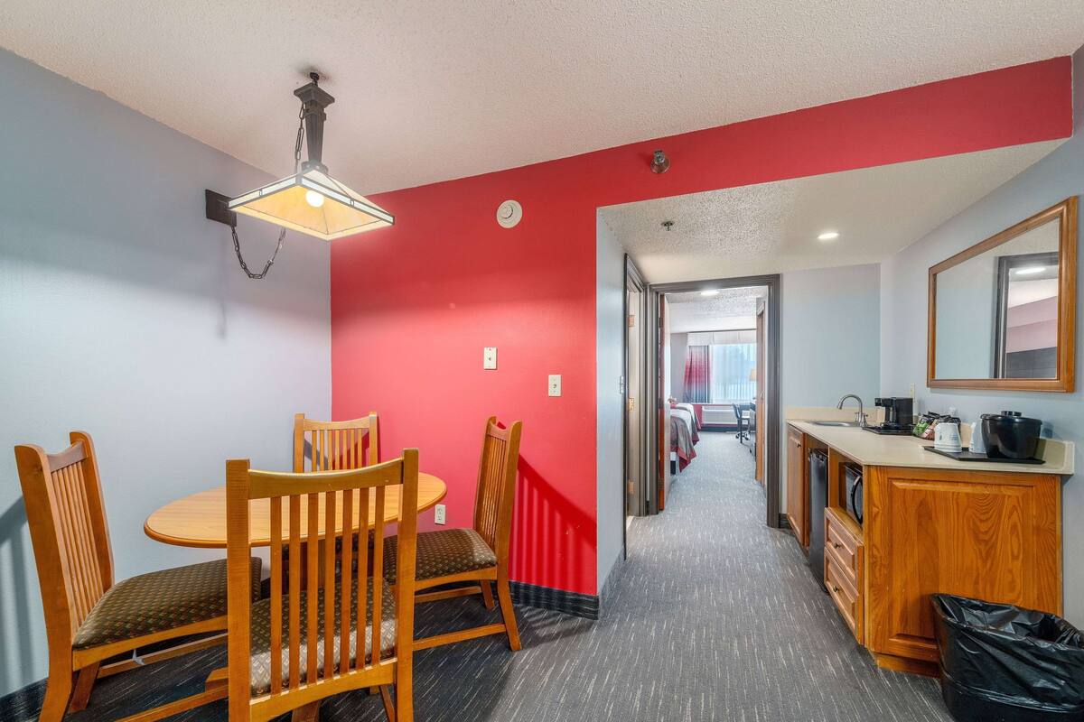 A hotel room with a dining table, chairs, a kitchen area, and a hallway leading to another room.