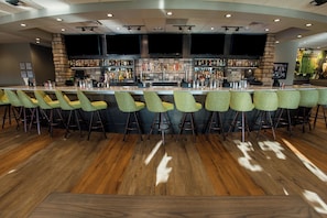 A bar area with a long counter, multiple stools, and a variety of bottles and glasses. - Holiday Inn Hotel & Suites Des Moines - Northwest by IHG (Urbandale)