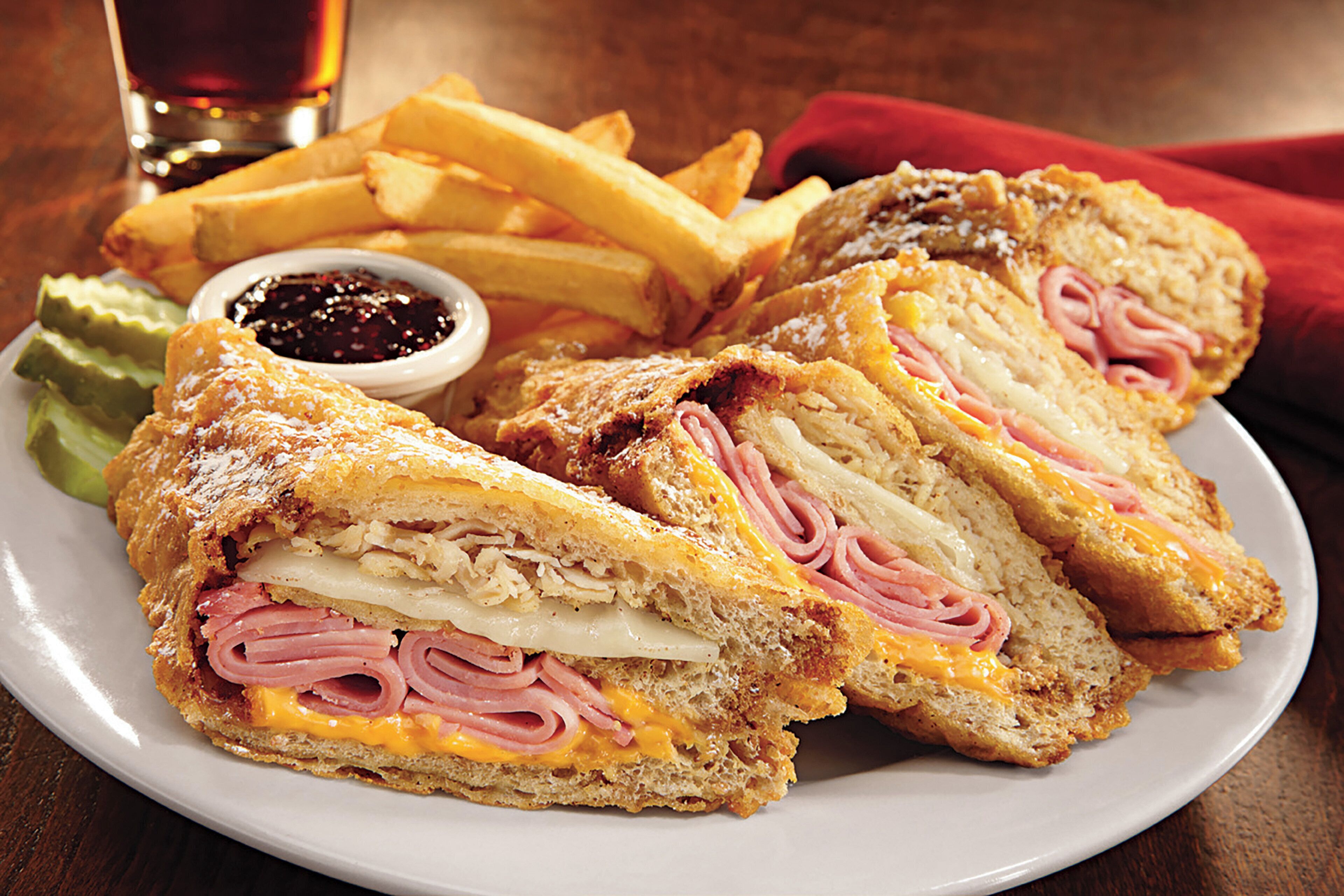 A plate with a sandwich cut in half, french fries, a small bowl of sauce, and a glass of a dark beverage.