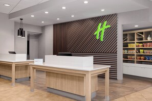 A reception area with two reception desks, a modern light fixture, a wall with a geometric pattern, and a shelf with snacks. - Holiday Inn Hotel & Suites Des Moines - Northwest by IHG (Urbandale)