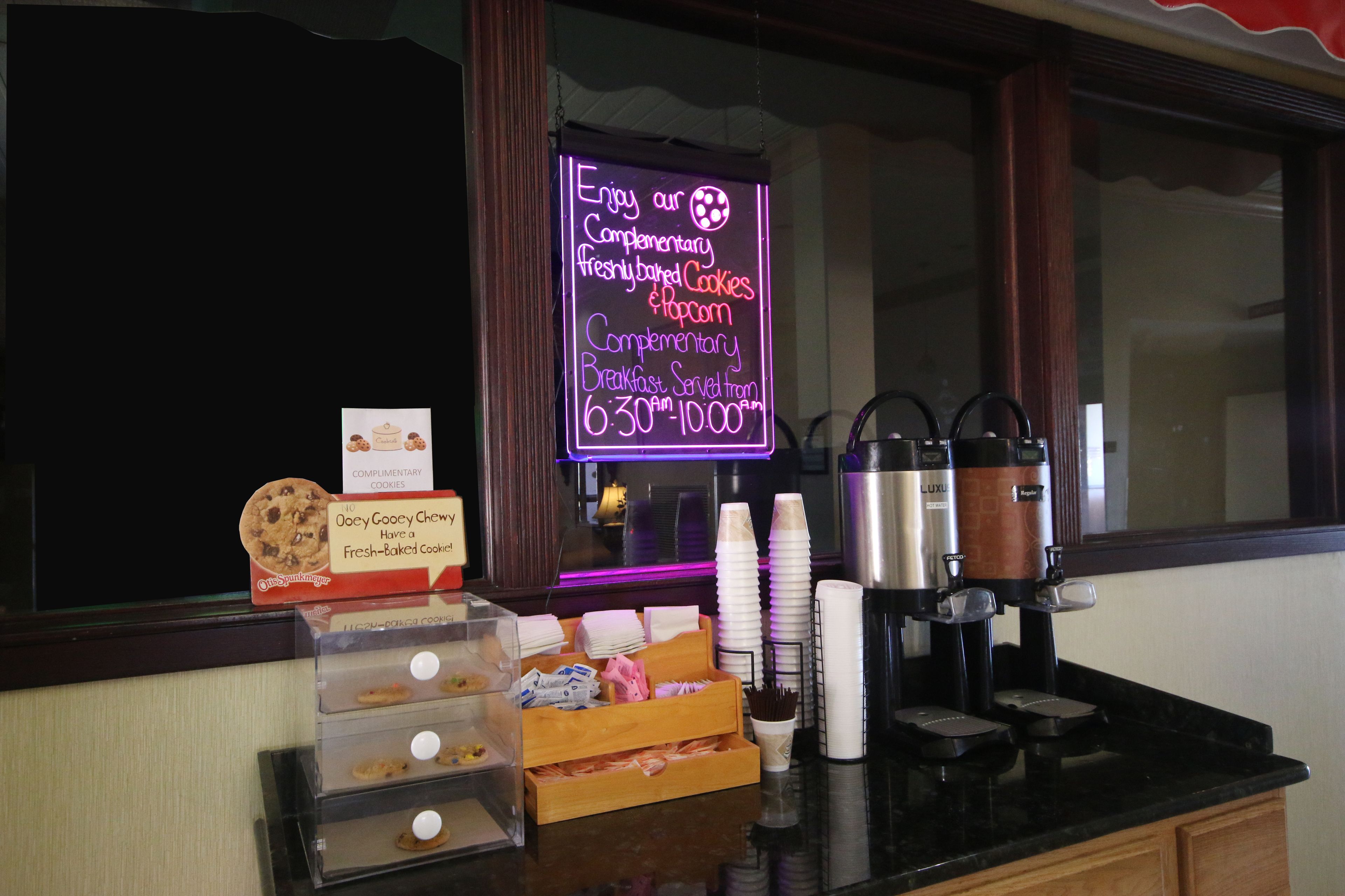 A breakfast bar with a neon sign advertising complimentary freshly baked cookies and popcorn, a coffee machine, and a display of cookies and pastries.
