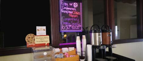 A breakfast bar with a neon sign advertising complimentary freshly baked cookies and popcorn, a coffee machine, and a display of cookies and pastries.