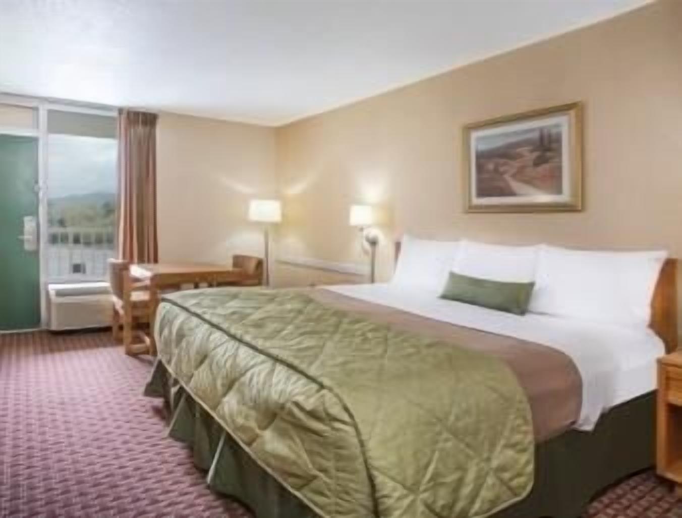 Quarto de hotel com cama, escrivaninha, cadeira, abajur e janela com cortinas.