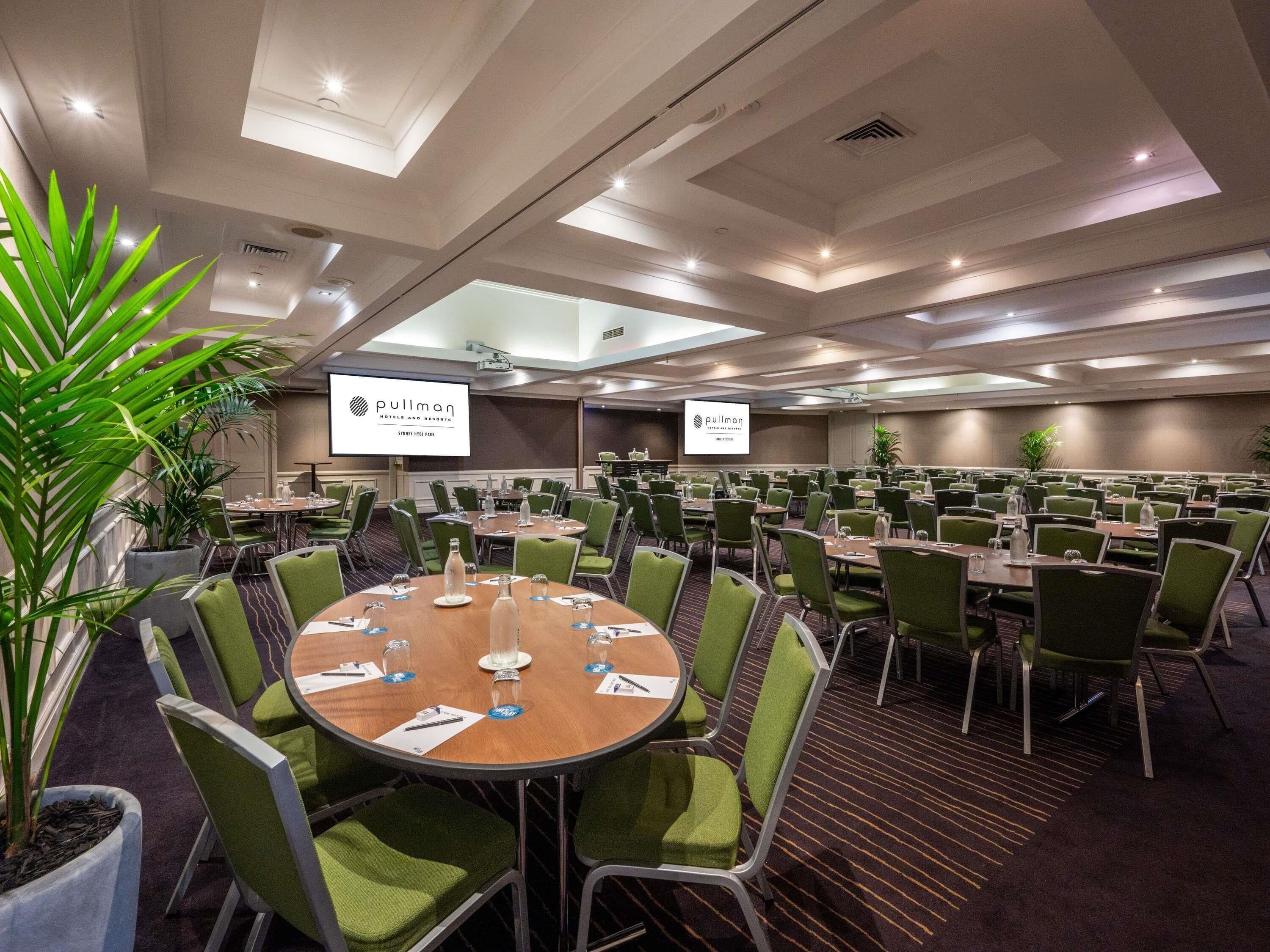 Un salón de conferencias con varias mesas y sillas, equipado con botellas de agua y placas de identificación, y una pantalla grande que muestra el nombre del hotel.