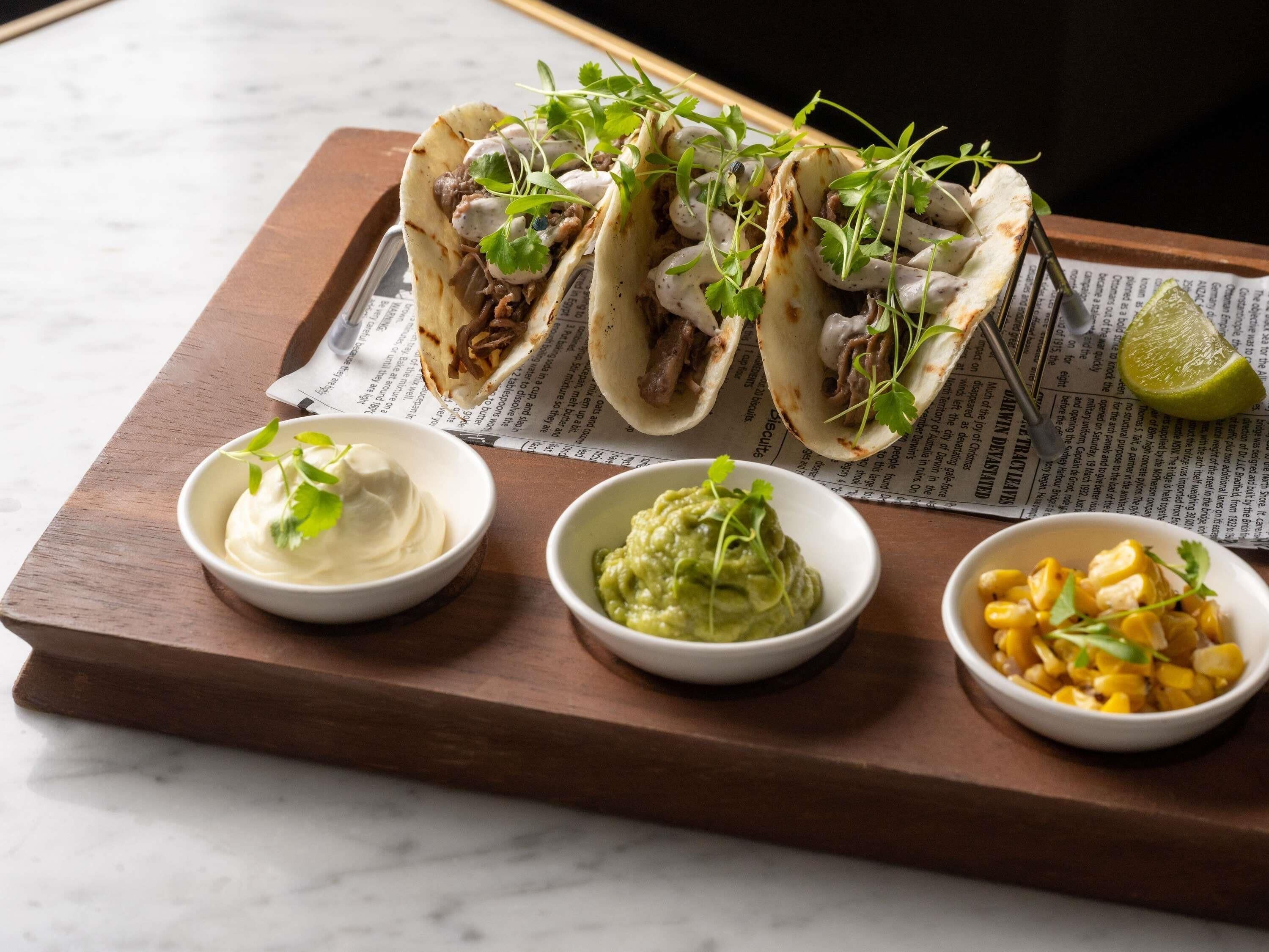 Drei Tacos mit Belag, zwei Soßen-Schalen und eine Limette auf einem Holzboden.