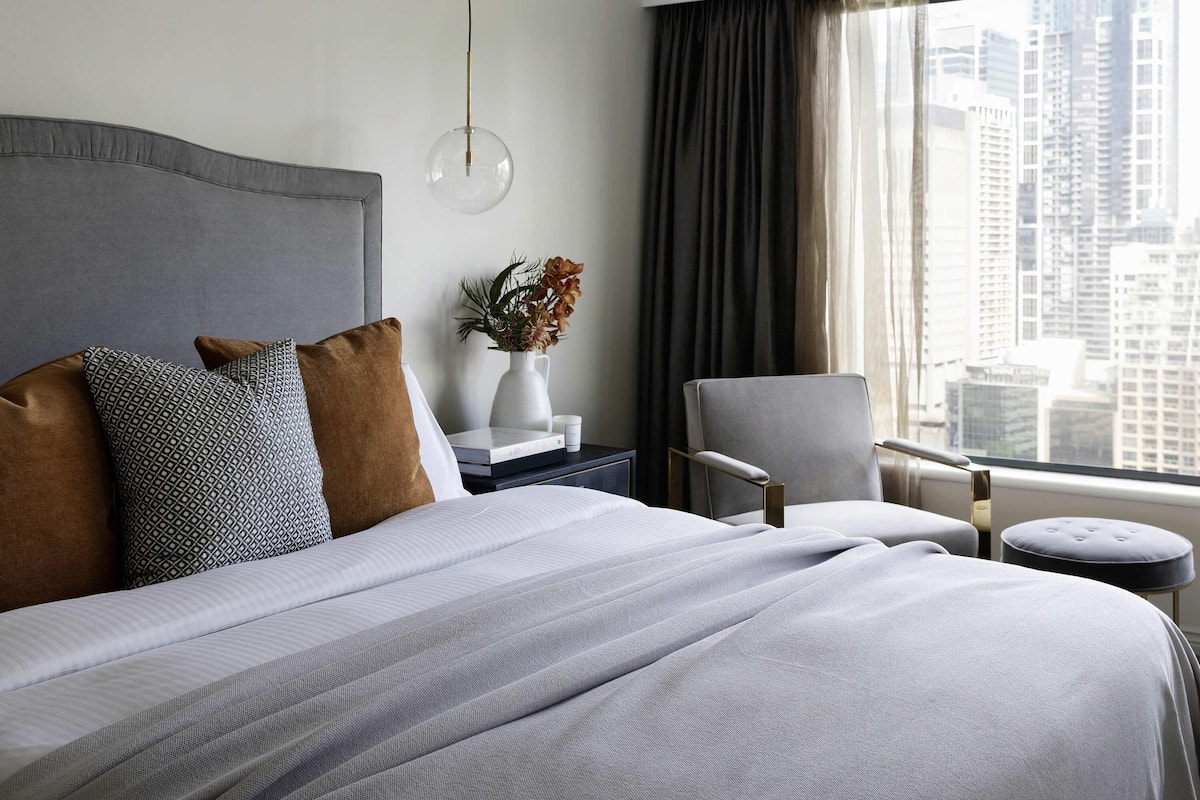 Een moderne hotelkamer met een groot bed, een nachtkastje, een stoel en uitzicht op hoge gebouwen.