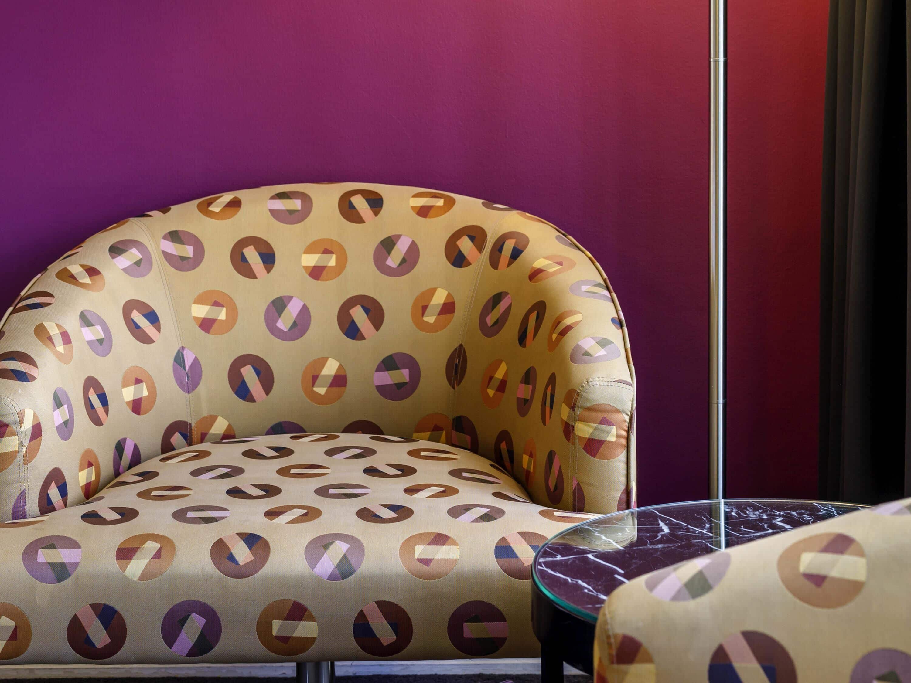 A room with a purple wall, a patterned armchair, and a glass table.
