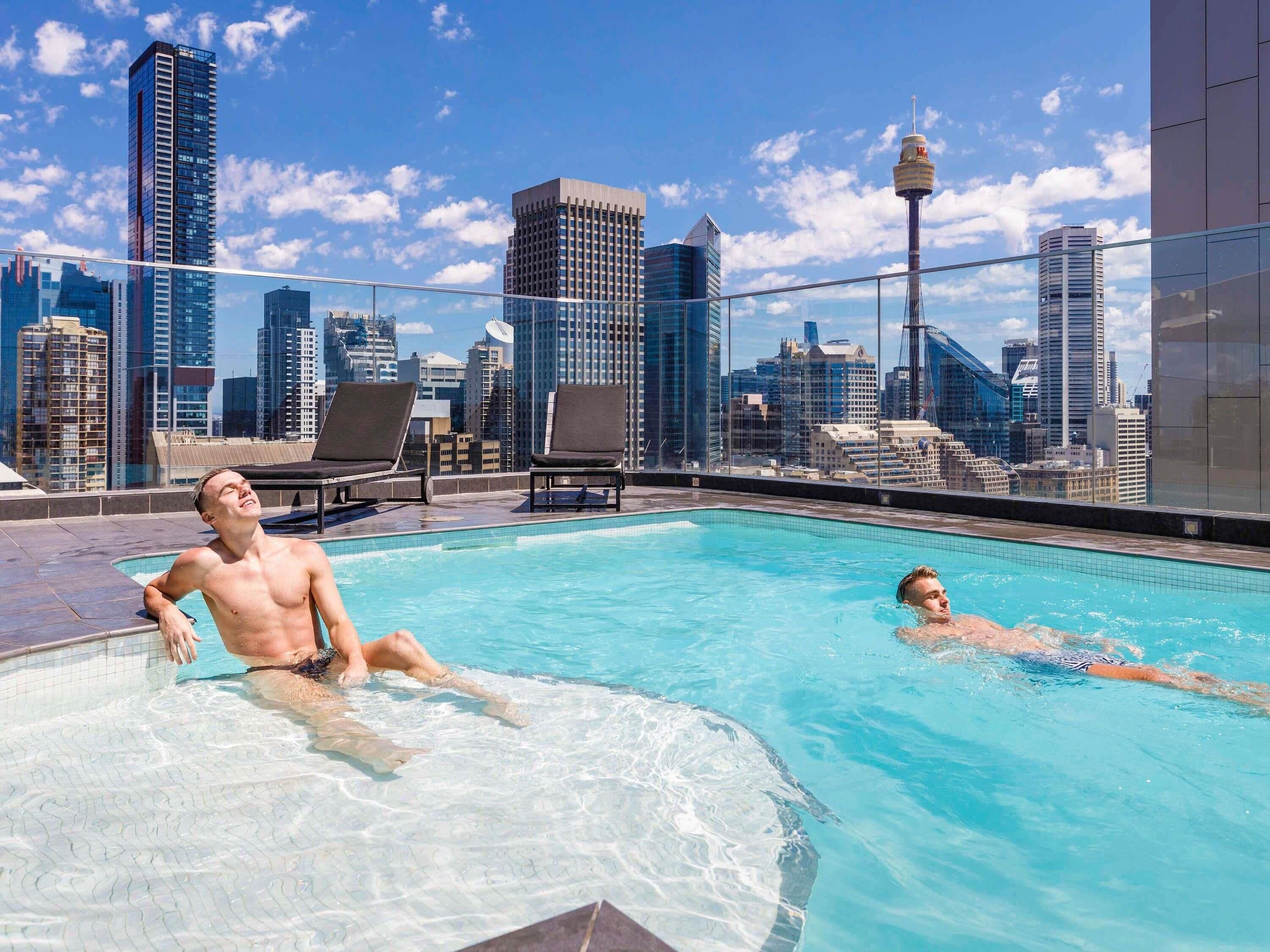A rooftop pool with two lounge chairs and a city skyline in the background.