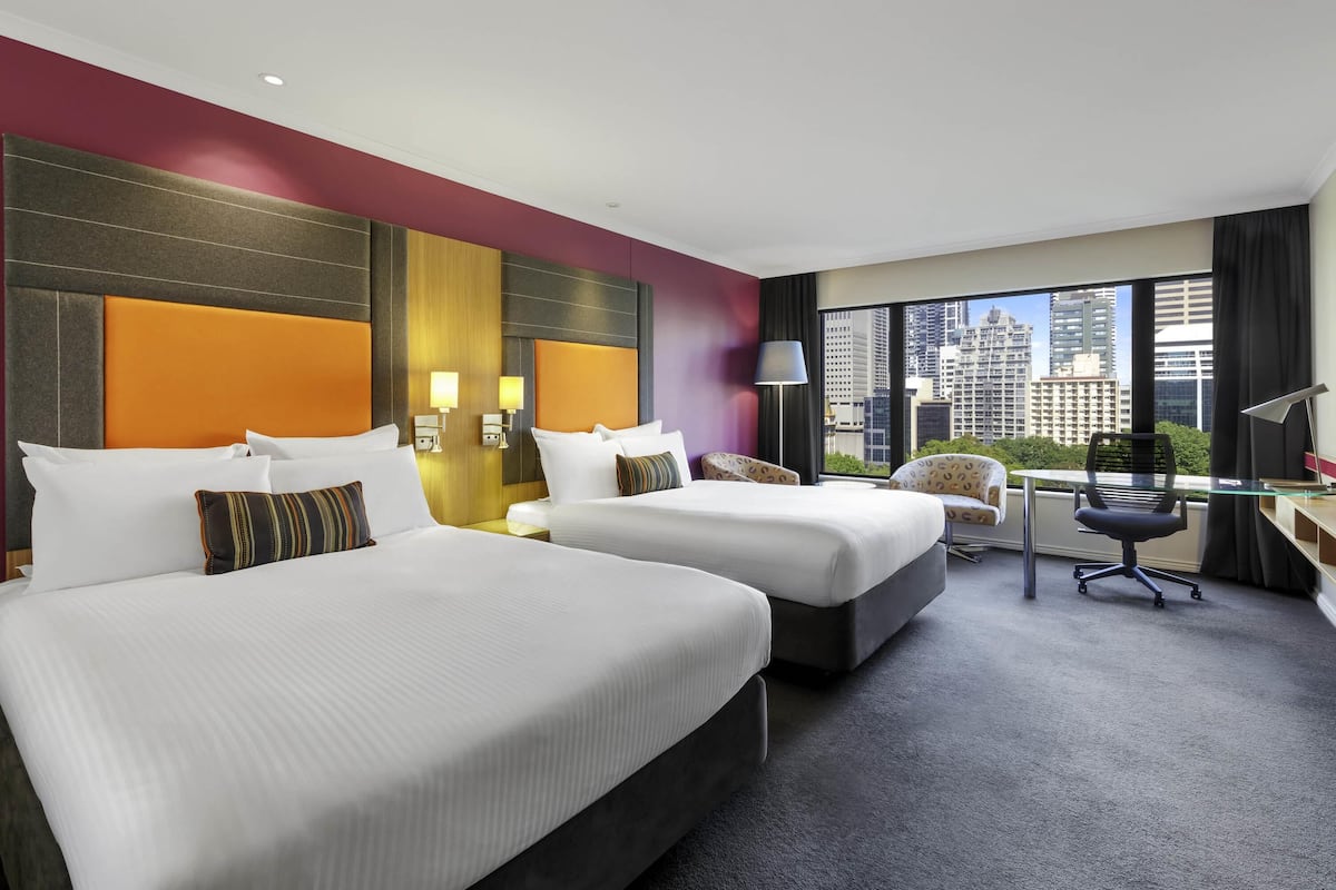 Een hotelkamer met twee bedden, een bureau, een stoel en een groot raam met uitzicht over de stad.