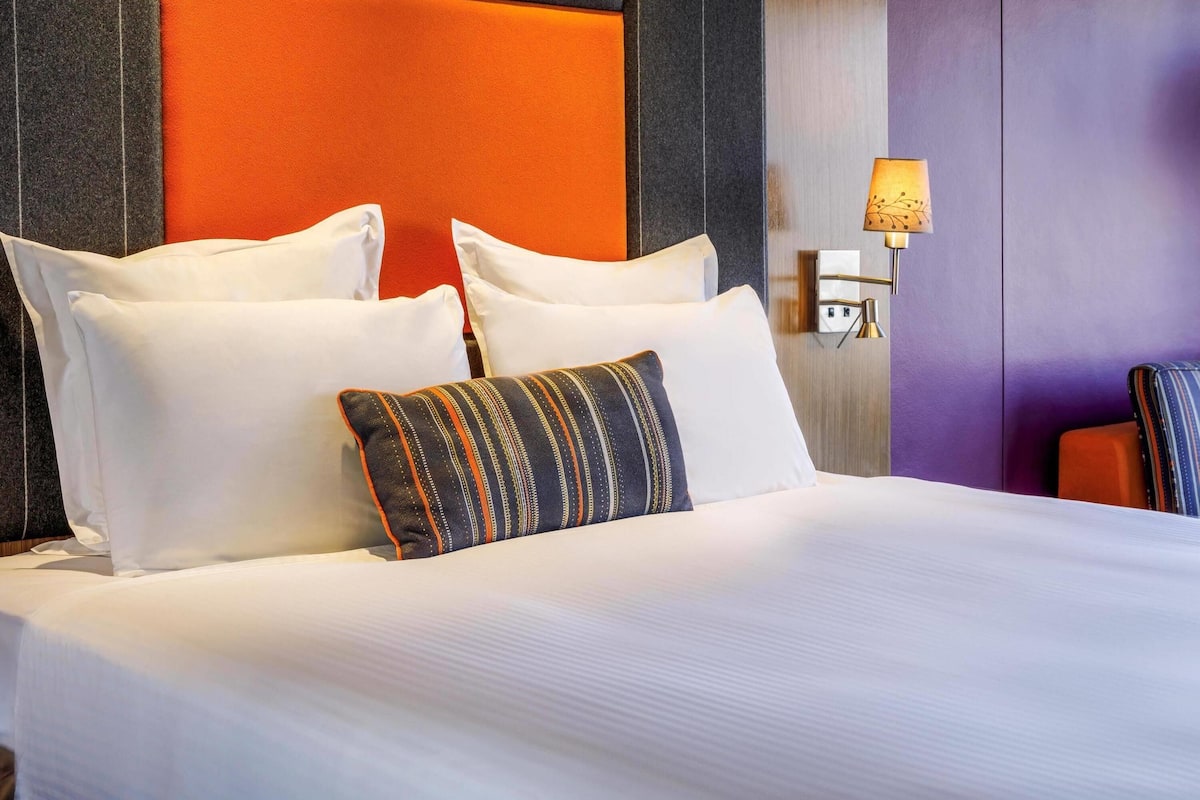 Een hotelkamer met een groot bed, witte kussens, een gestreept kussen, een wandlamp en een paars accentmuur.