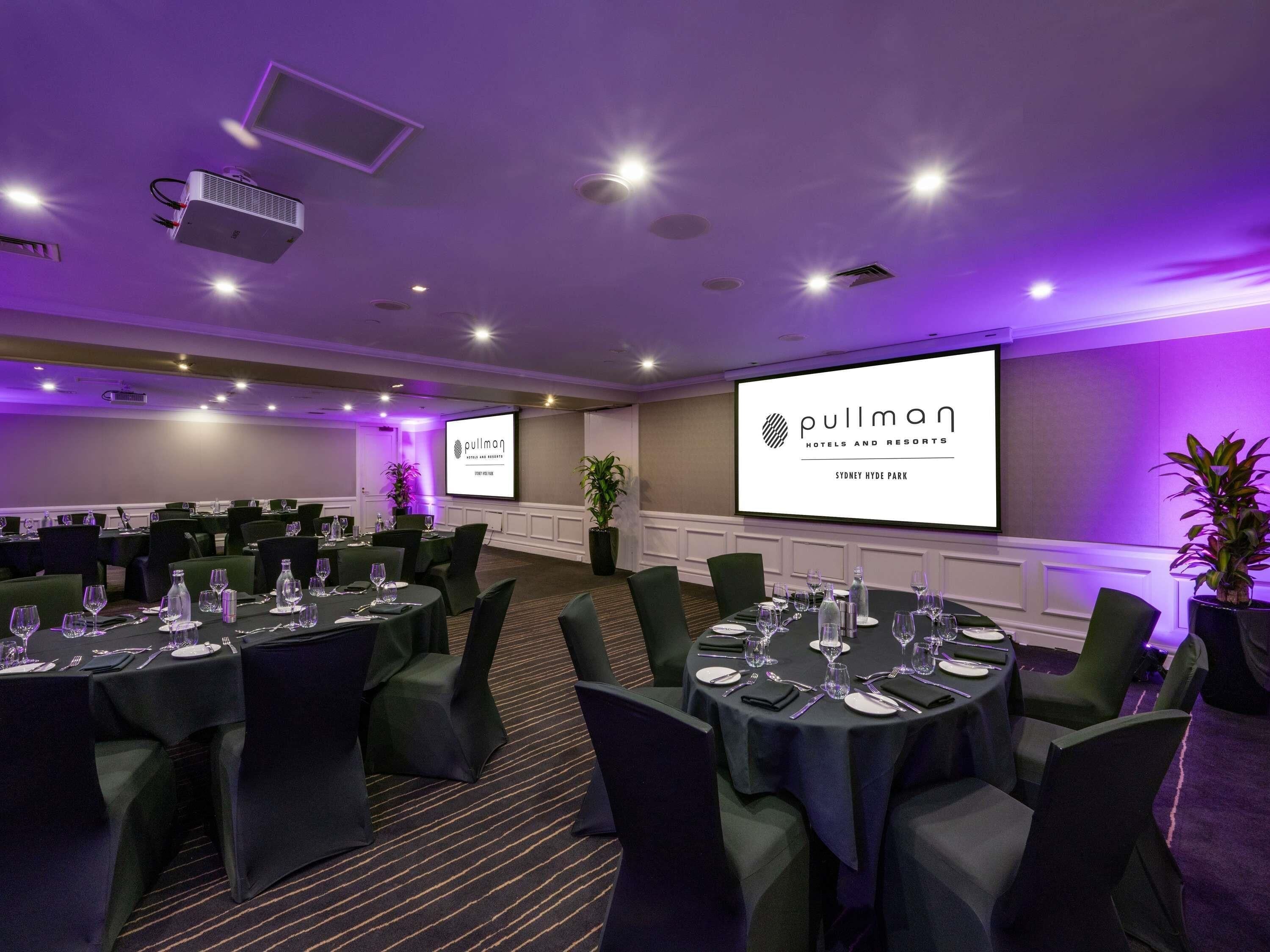 A conference room with multiple tables set for a meeting, a large screen displaying the hotel name, and a projector in the ceiling.