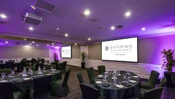 A conference room with multiple tables set for a meeting, a large screen displaying the hotel name, and a projector in the ceiling.