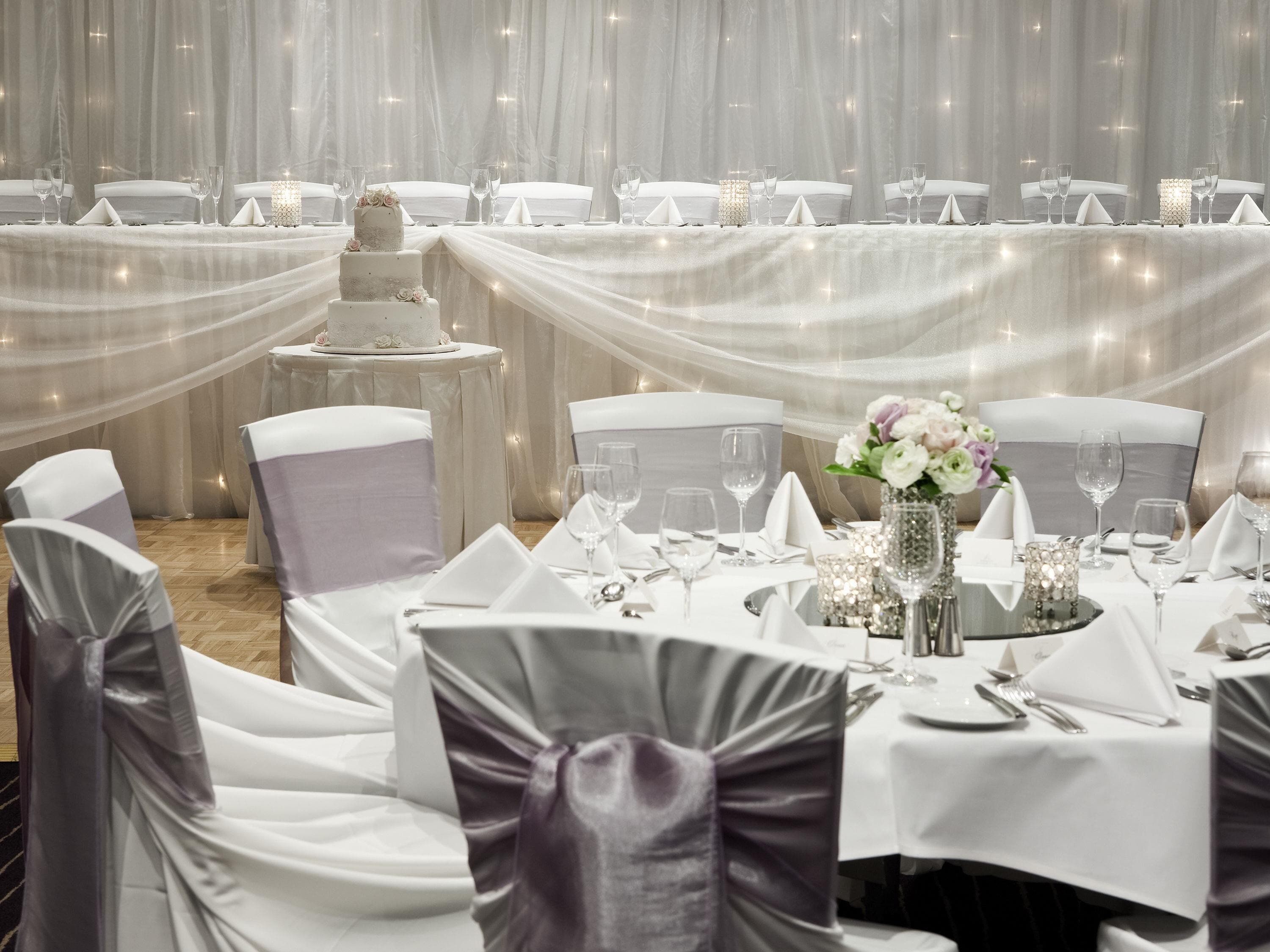 A banquet hall with round tables, chairs, and a tiered wedding cake.
