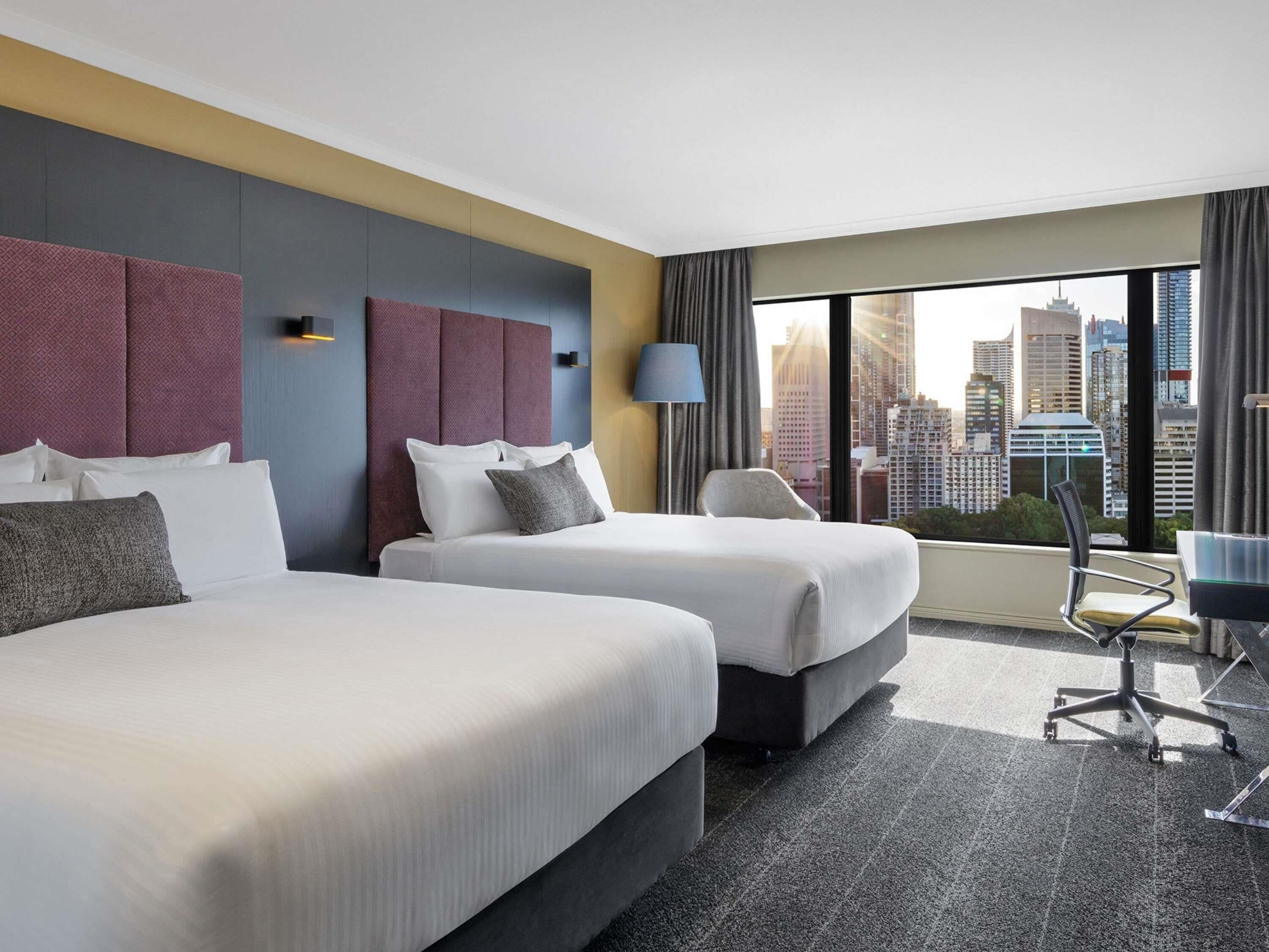 A hotel room with two beds, a desk, and a view of the city skyline.