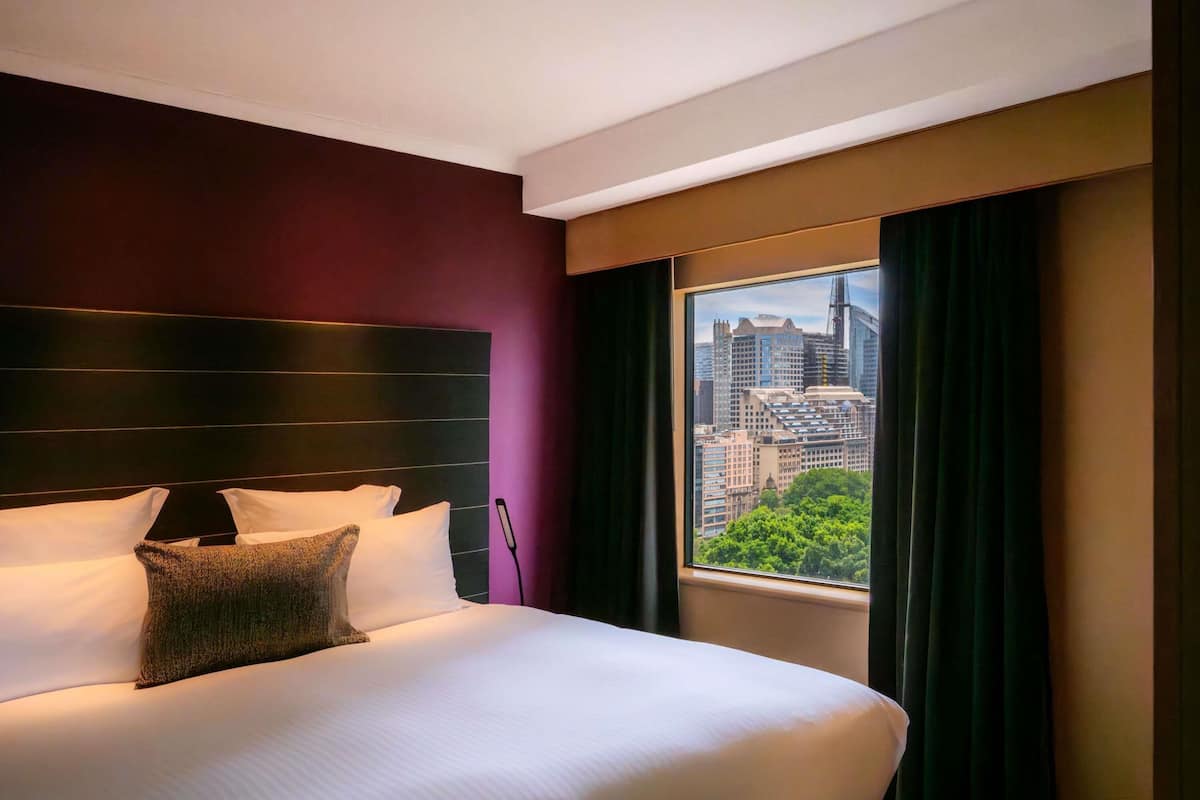 Een hotelkamer met een groot bed, een hoofdbord, een raam met gordijnen en uitzicht op de stad.