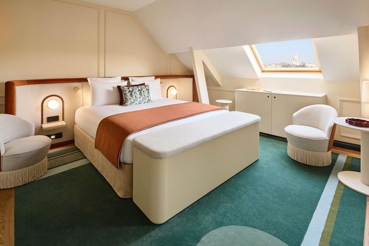 A hotel room with a bed, bedside tables, a desk, a chair, and a view through a skylight.