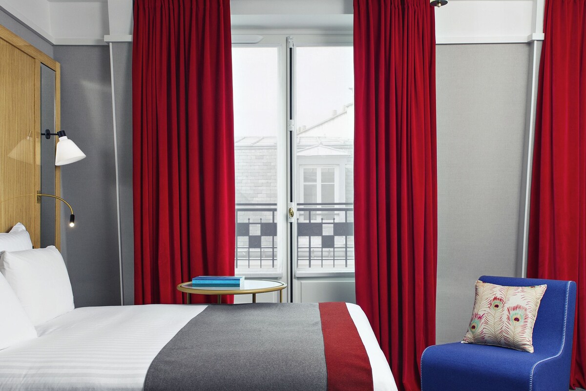 A modern hotel room with a large bed, a blue armchair, a small table with books, and a view through a glass door.
