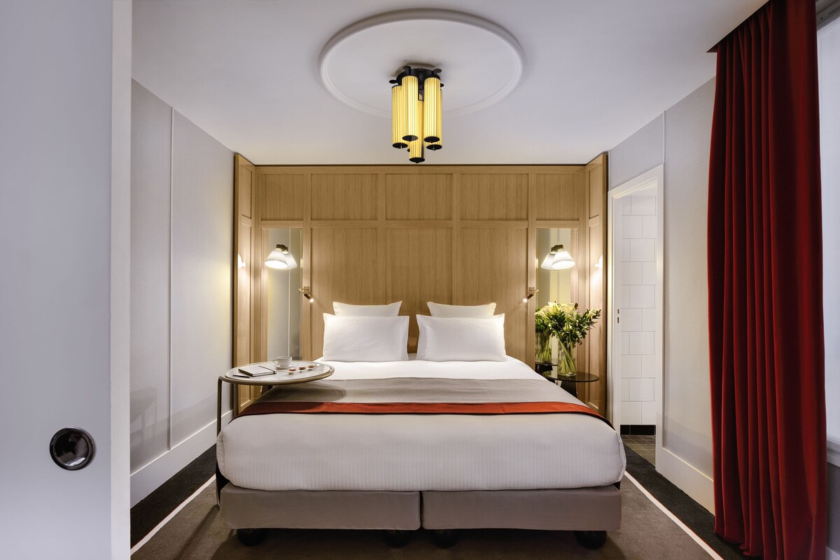 A hotel room with a large bed, two bedside tables, a round mirror, and a ceiling light fixture.