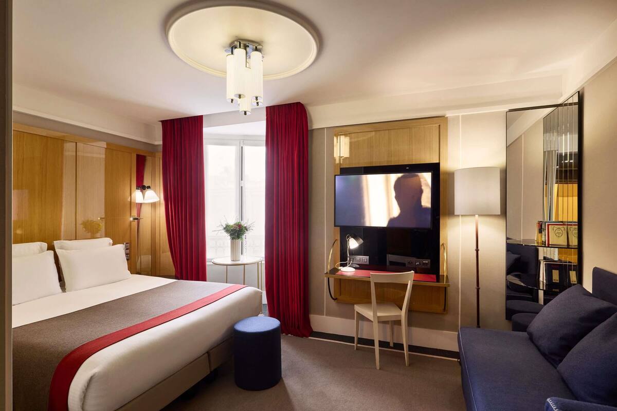 A hotel room with a bed, a desk with a computer, a TV, a chair, a sofa, and a small table with a plant.