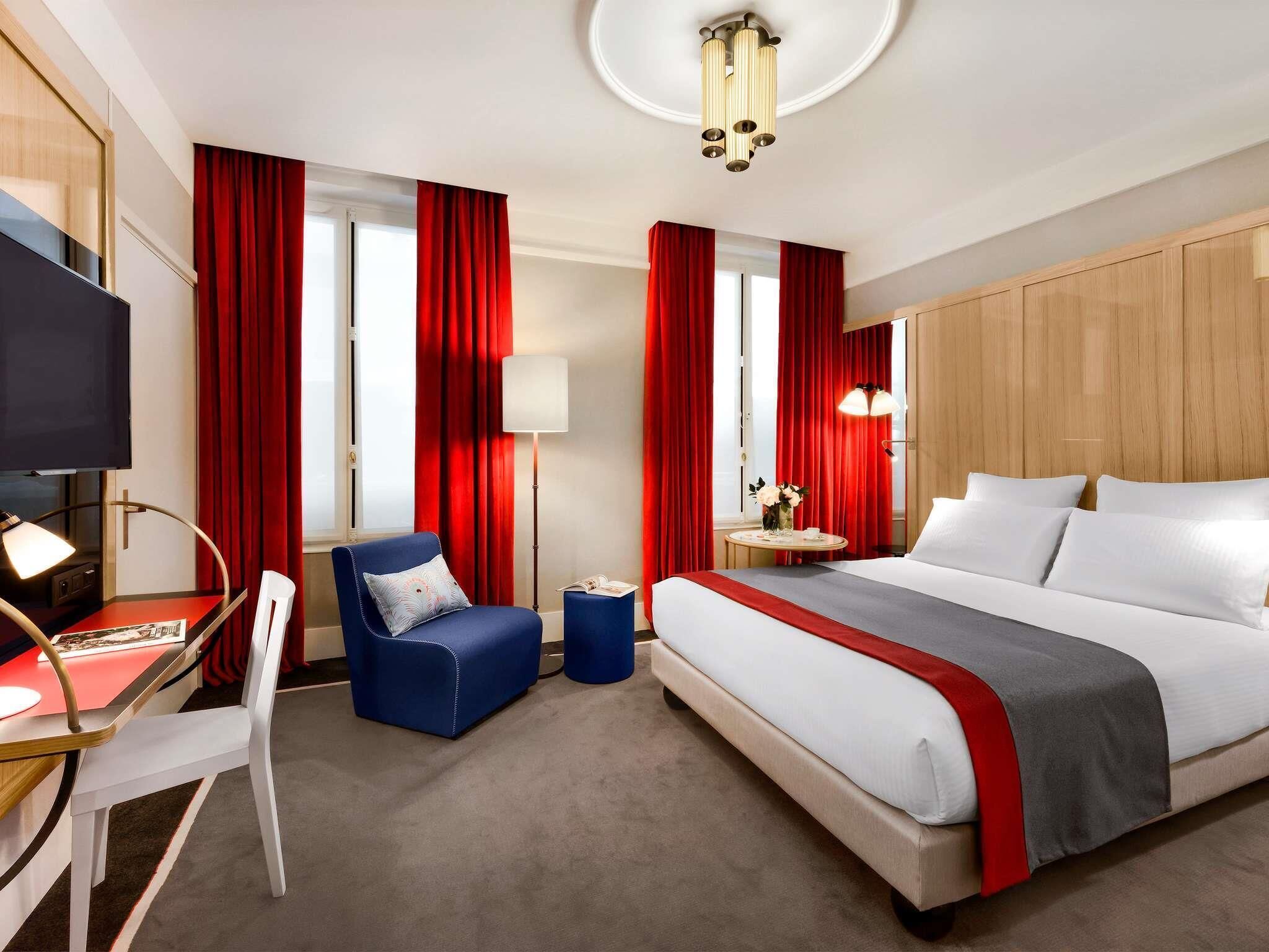 A hotel room with a bed, a desk with a chair, a television, and red curtains.