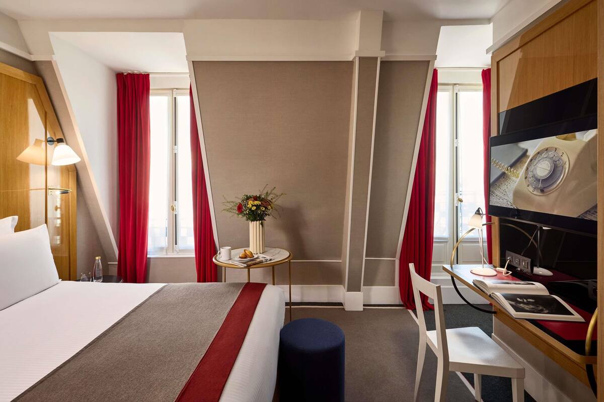 A hotel room with a bed, a desk, a chair, a small table with a vase of flowers, and a television.