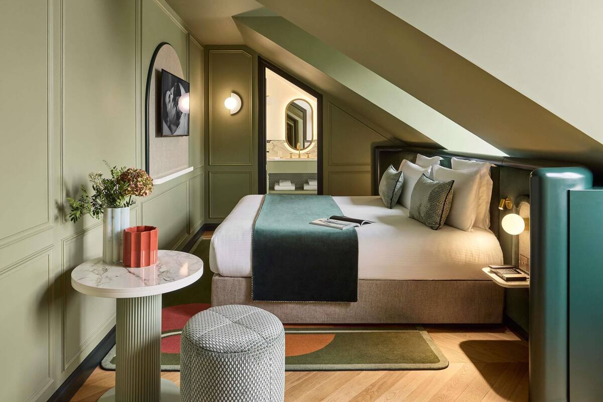 A modern bedroom with a large bed, a bedside table, a small round ottoman, and a nightstand with a lamp.