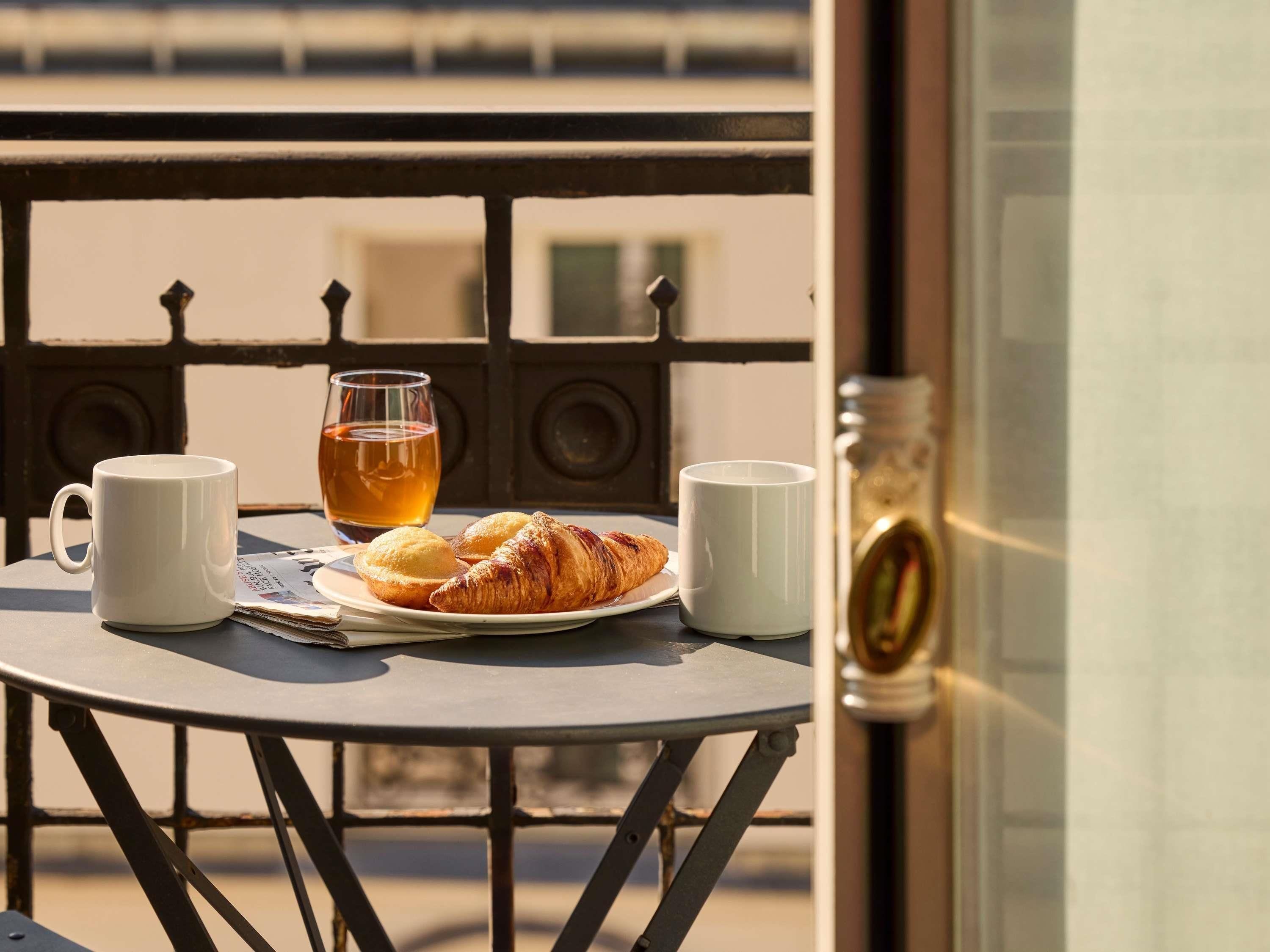 Ein Tisch mit einer Teetasse, zwei weißen Tassen und einem Teller mit Croissants auf einem Balkon.