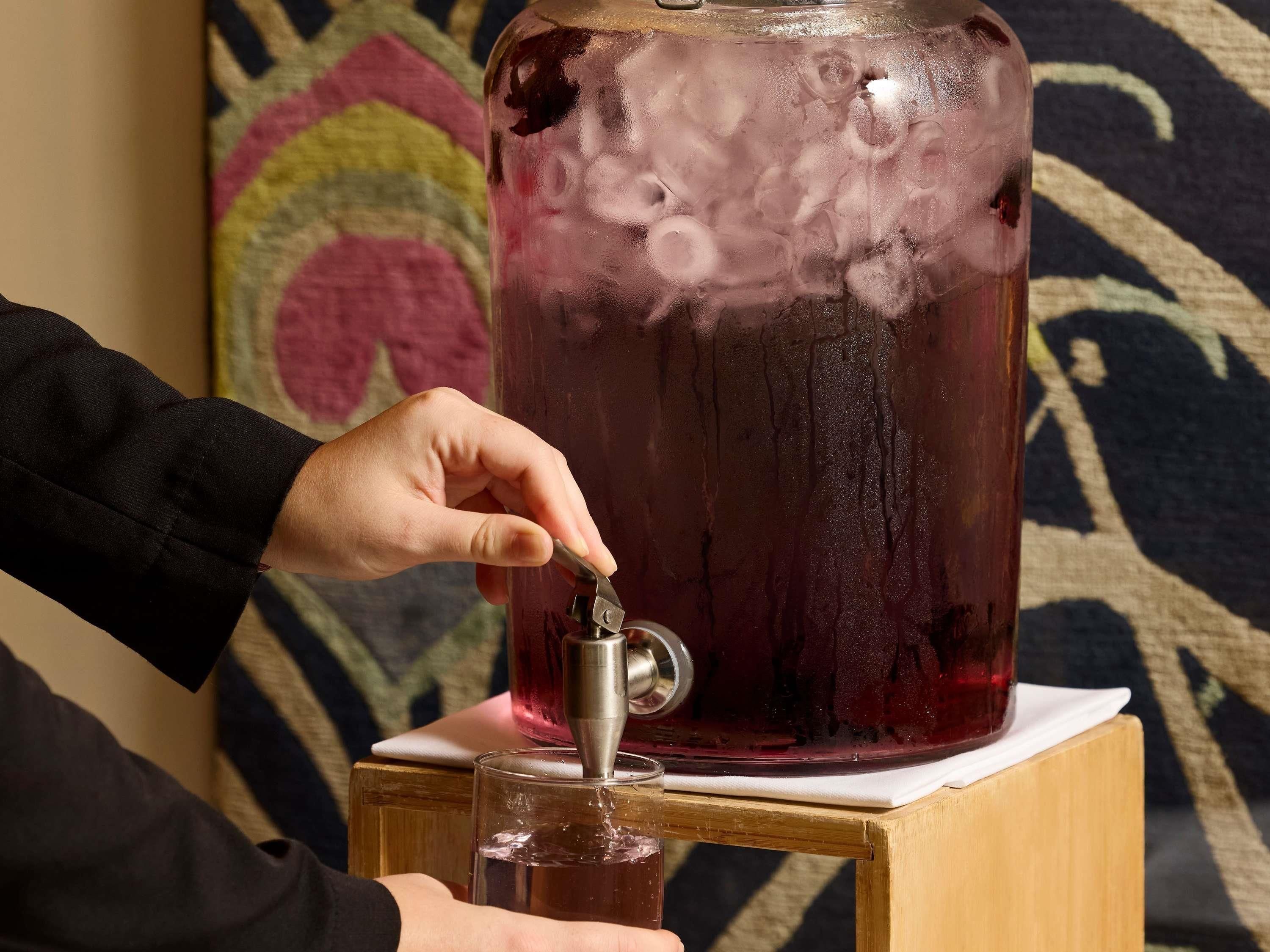A hand operating a tap on a large, clear container filled with a dark liquid, possibly for serving.
