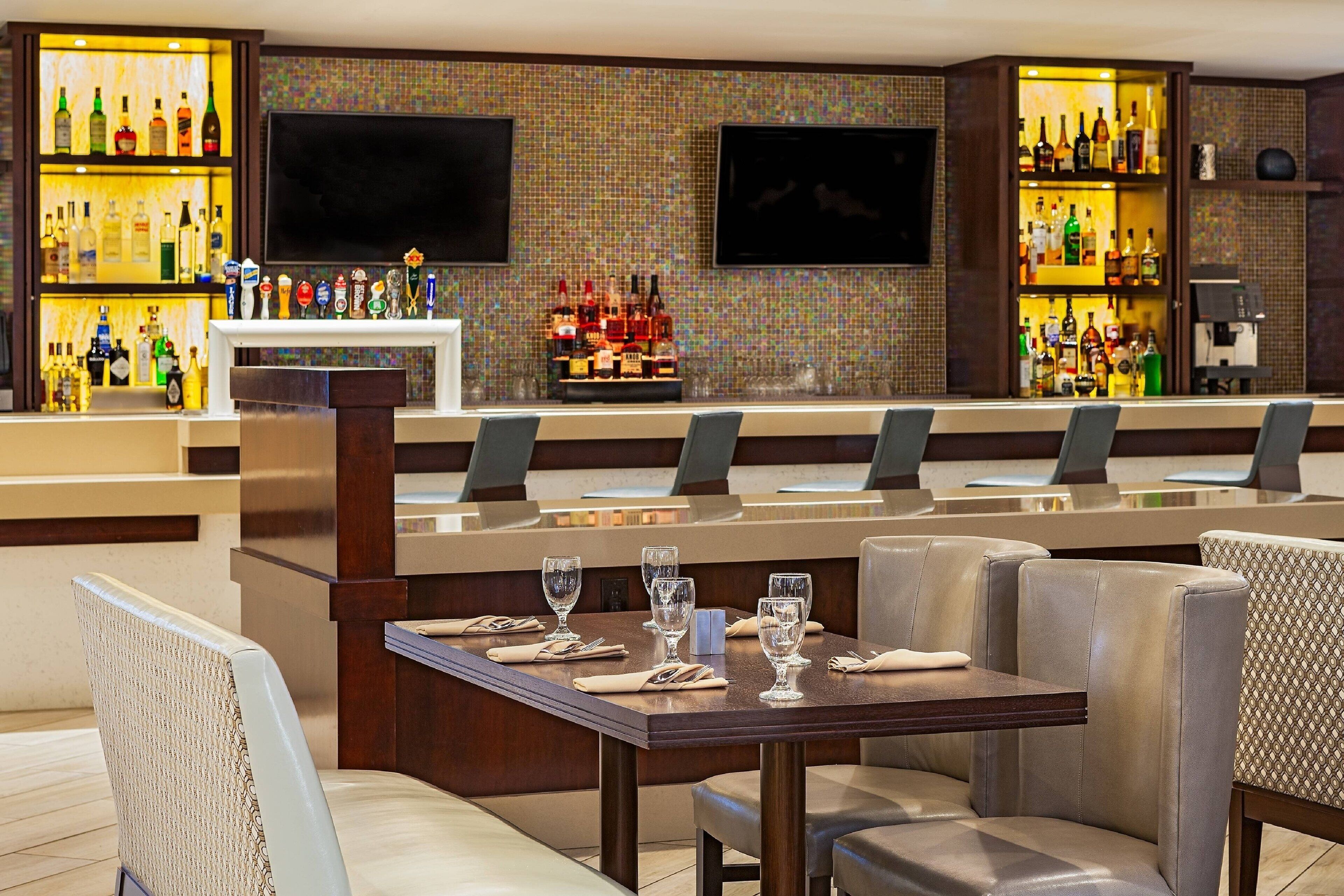 A bar area with multiple seating options, a television mounted on the wall, and shelves stocked with various bottles.