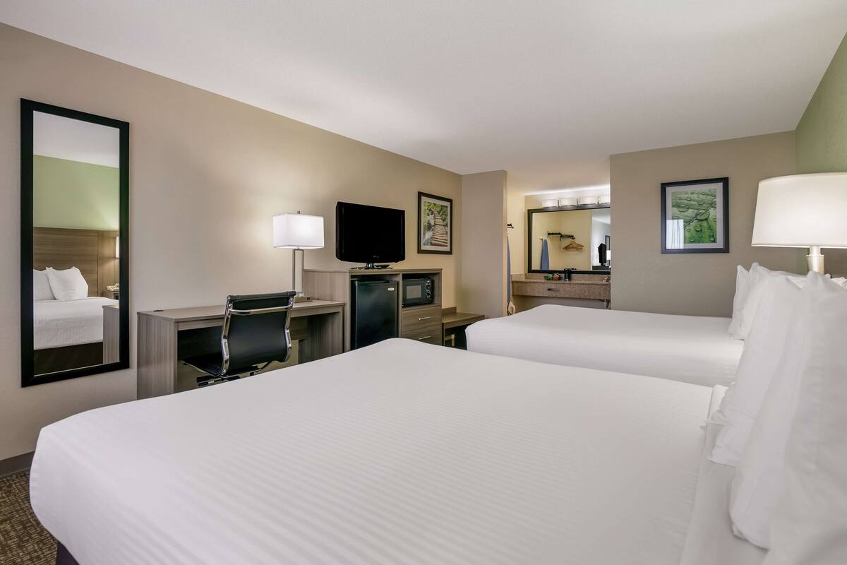 A hotel room with two beds, a desk with a chair, a television, a mirror, and a small kitchenette.