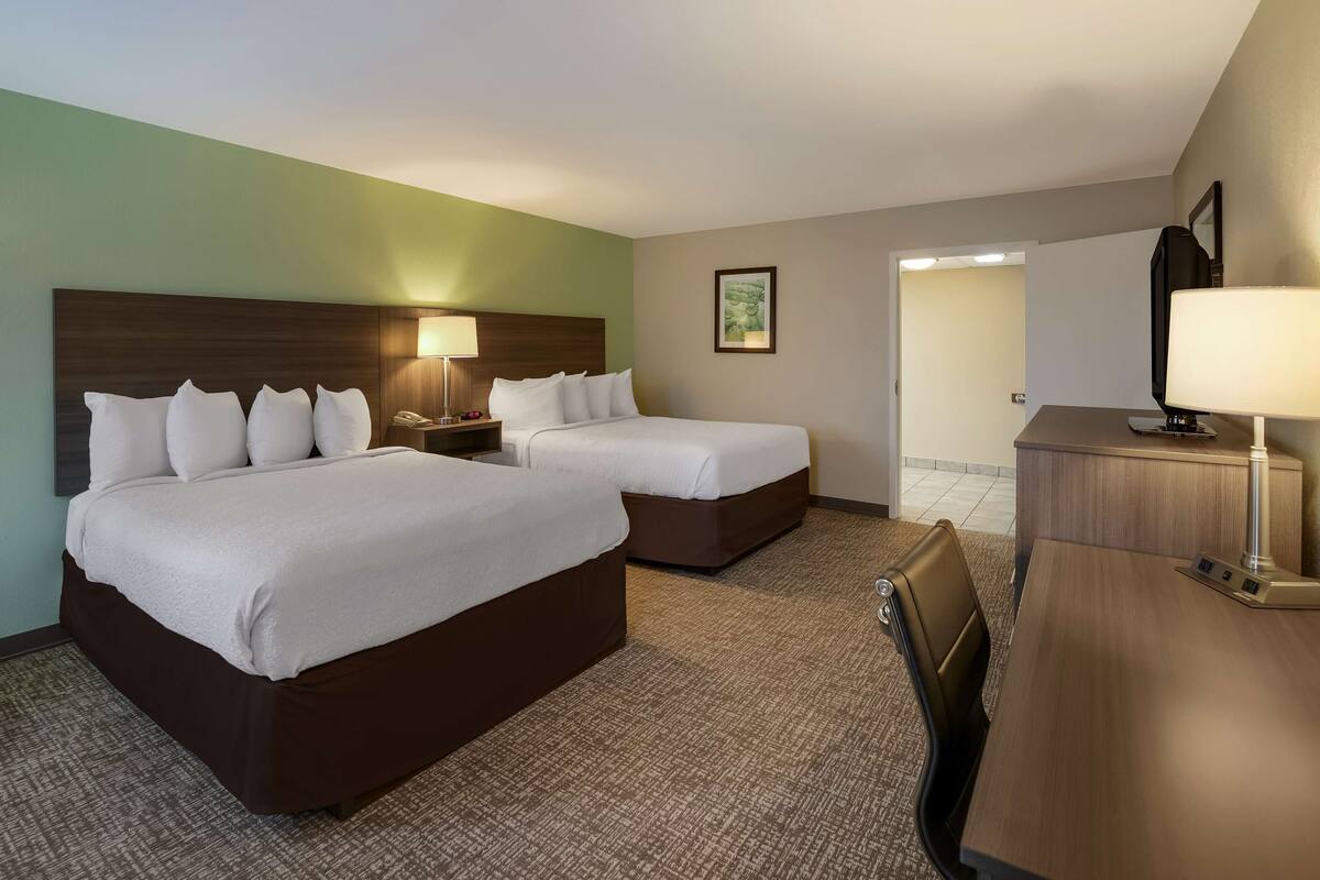 A hotel room with two beds, a desk, a chair, and a lamp.