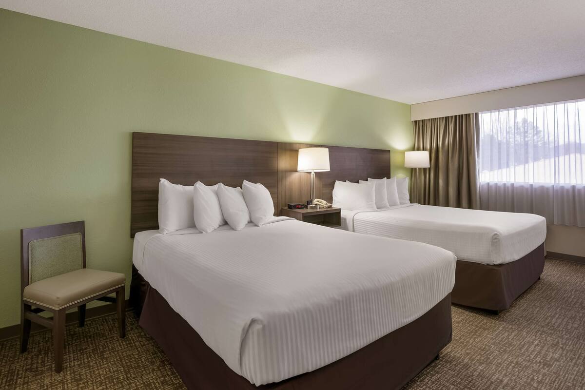 A hotel room with two beds, a chair, a nightstand with a lamp, and a window with curtains.