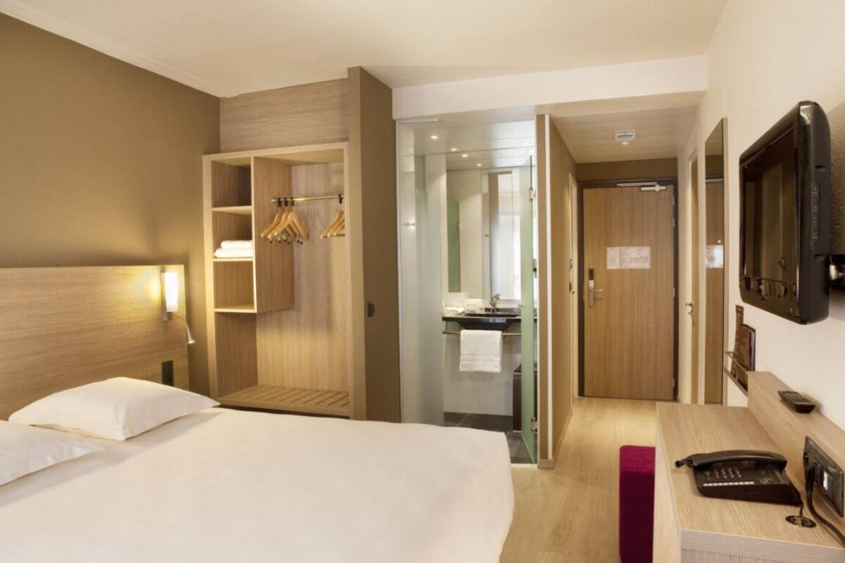 Habitación de hotel con cama, escritorio con teléfono, televisión, armario y baño visible a través de una puerta abierta.
