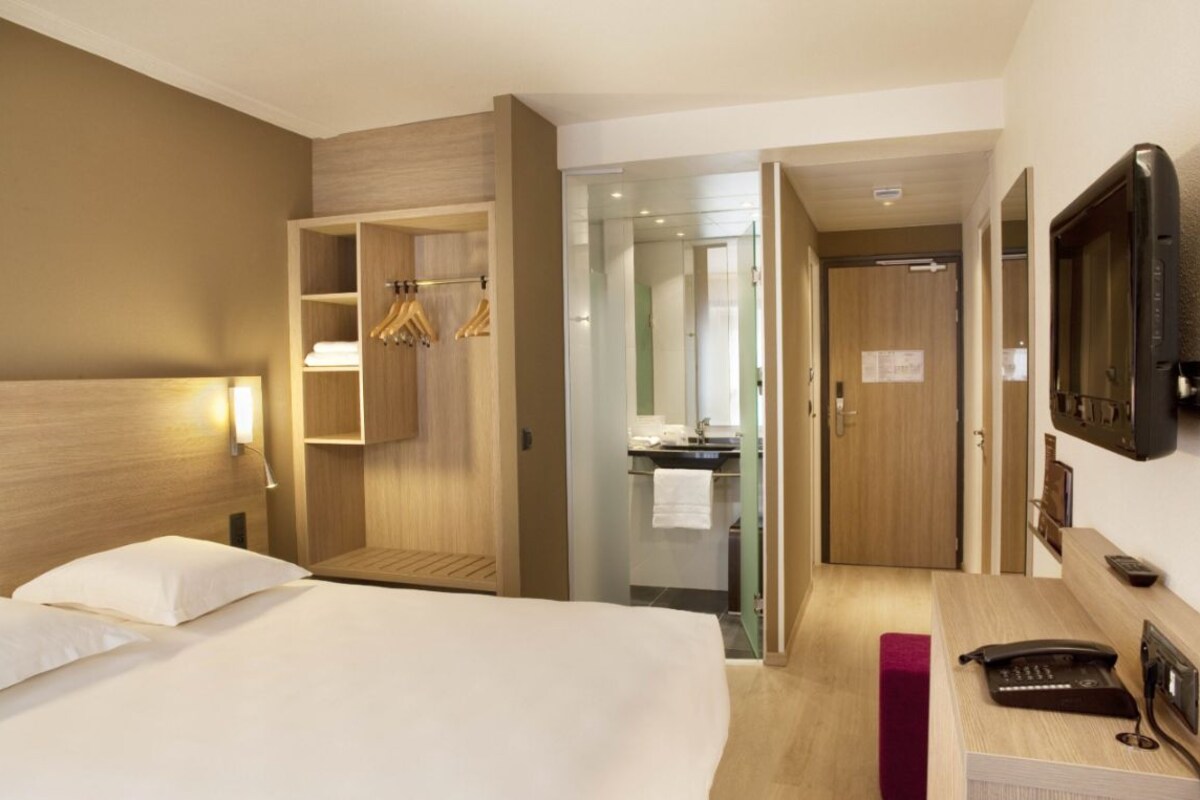 Quarto de hotel com uma cama, uma escrivaninha com telefone, televisão, guarda-roupa e um banheiro visível através de uma porta aberta.