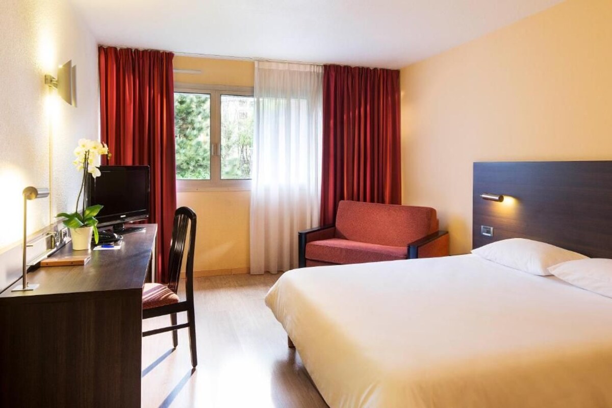 Una habitación de hotel con cama, escritorio con televisor, silla y ventana con cortinas.