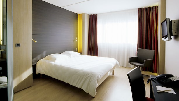 A hotel room with a bed, a desk with a chair, a television, and a window with curtains.