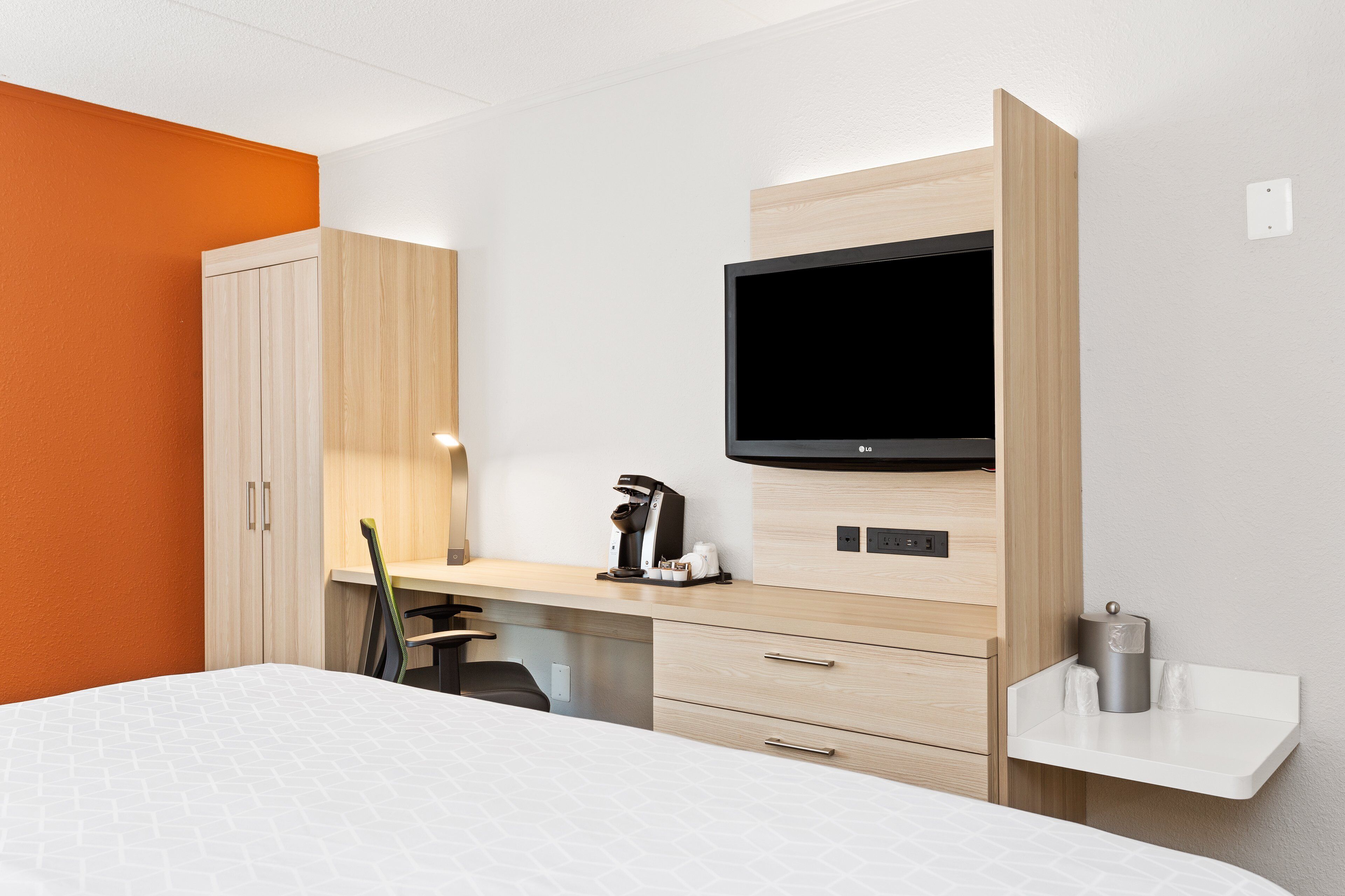 Una habitación de hotel con una cama, un escritorio con cafetera, un televisor y un estante pequeño.