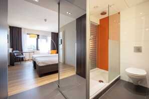 Ein modernes Hotelzimmer mit Bett, Schreibtisch, Stuhl und einer Dusche mit Glastüren.