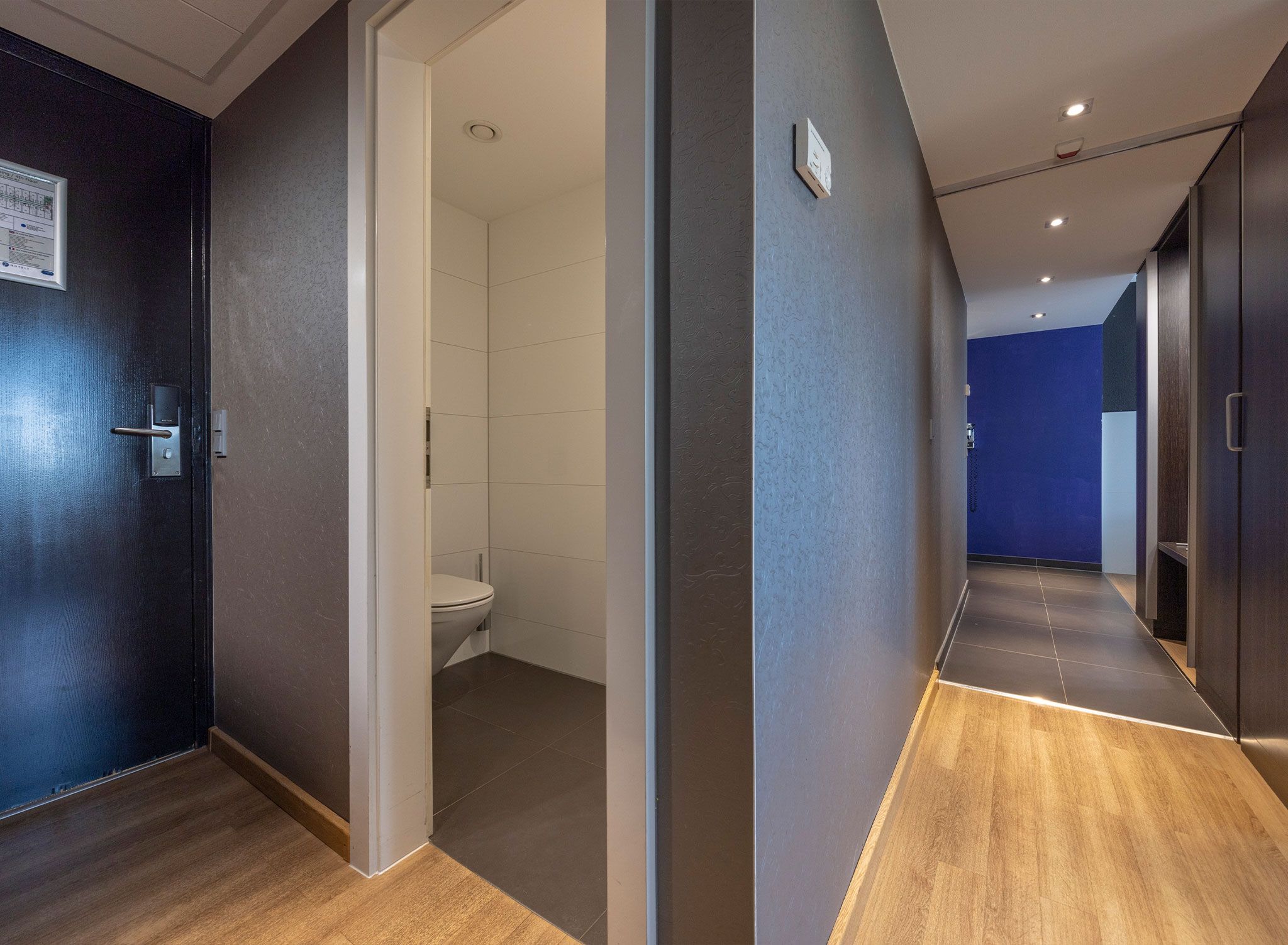 Un pasillo moderno de hotel con piso de madera, un baño a la vista con inodoro y una pared azul de acento.